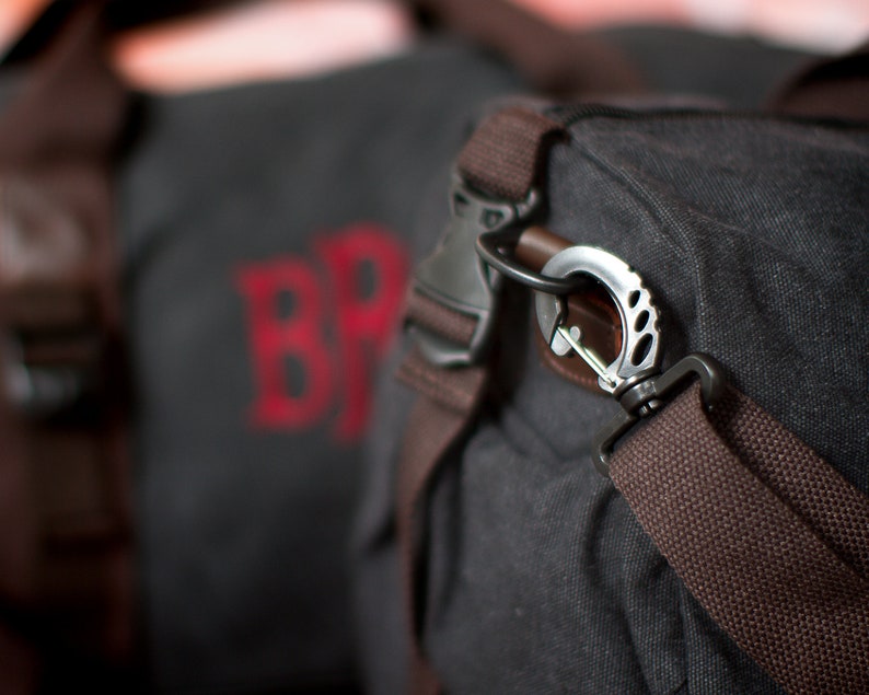 K&ouml;nnte beinhalten: Eine schwarze Canvas-Reisetasche mit braunen Riemen und einem silbernen Metallhaken. Die Tasche hat ein rotes gesticktes Logo mit der Aufschrift "BR".