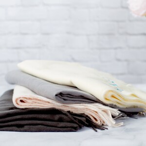 May include: A stack of four scarves in different shades of gray, beige, and pink. The scarves are folded and arranged in a pile on a white surface.