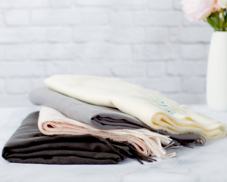 May include: A stack of four scarves in different shades of gray, beige, and pink. The scarves are folded and arranged in a pile on a white surface.
