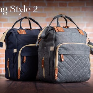 May include: Two backpacks labeled Bag Style 2, one navy blue and one heather gray. Both feature top handles, beige zippers, brown leather-like accents, and a quilted front pocket design. A small matching circular pouch is attached to the gray bag.