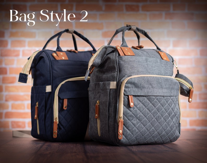 May include: Two backpacks labeled Bag Style 2, one navy blue and one heather gray. Both feature top handles, beige zippers, brown leather-like accents, and a quilted front pocket design. A small matching circular pouch is attached to the gray bag.