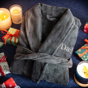 May include: A dark gray plush robe with a white embroidered "Dad" on the front. The robe is tied at the waist and has a shawl collar. The robe is surrounded by wrapped gifts and candles.