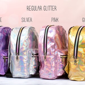 May include: Four mini backpacks in different colors: purple, silver, pink, and gold. The backpacks have a holographic finish and feature a zipper closure. The text "REGULAR GLITTER" is displayed above the backpacks, and the colors are labeled below each backpack.