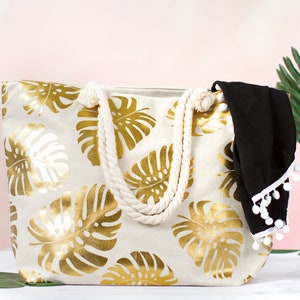 May include: A white canvas tote bag with gold foil tropical leaf print and rope handles. The bag has a black fabric lining with white pom pom trim.