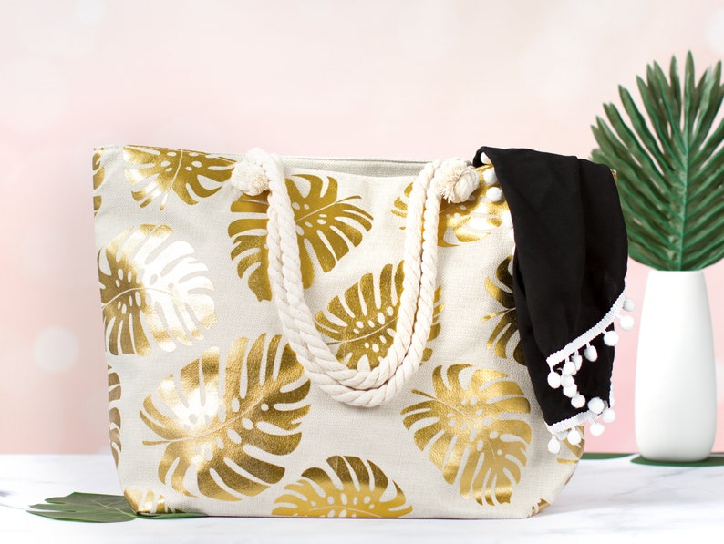 May include: A white canvas tote bag with gold foil tropical leaf print and rope handles. The bag has a black fabric lining with white pom pom trim.