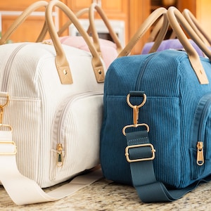 May include: Several corduroy travel bags in various colors, including white and blue, are displayed. Each bag has a zippered main compartment, a front pocket, and a detachable shoulder strap. The bags feature tan handles and gold-colored hardware.
