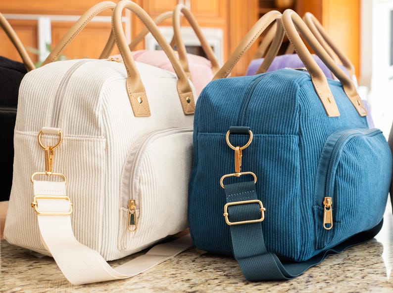 May include: Several corduroy travel bags in various colors, including white and blue, are displayed. Each bag has a zippered main compartment, a front pocket, and a detachable shoulder strap. The bags feature tan handles and gold-colored hardware.