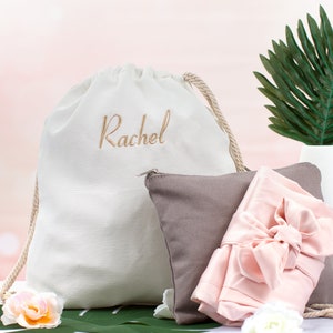 May include: A white drawstring bag with the name "Rachel" embroidered on it. The bag is next to a gray pillow and a pink satin ribbon.
