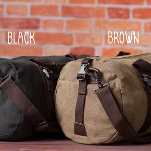 K&ouml;nnte beinhalten: Zwei Canvas-Reisetaschen, eine schwarze und eine braune, mit braunen Lederelementen und verstellbaren Riemen. Die Taschen sind mit "BLACK" und "BROWN" beschriftet.