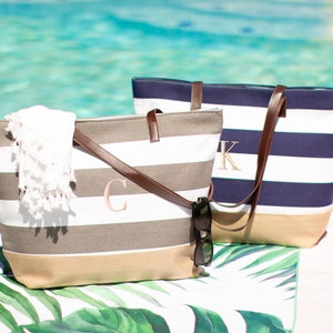 Puede incluir: Dos bolsas de lona a rayas con asas de cuero. La bolsa de la izquierda es gris y blanca con un monograma "C" rosa. La bolsa de la derecha es azul marino y blanca con un monograma "K" blanco.