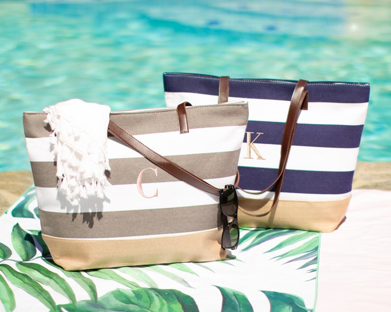 Puede incluir: Dos bolsas de lona a rayas con asas de cuero. La bolsa de la izquierda es gris y blanca con un monograma "C" rosa. La bolsa de la derecha es azul marino y blanca con un monograma "K" blanco.