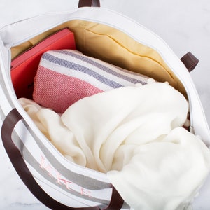 May include: A striped canvas tote bag with brown leather handles. The bag is filled with a white blanket, a red and white striped towel, and a red notebook.