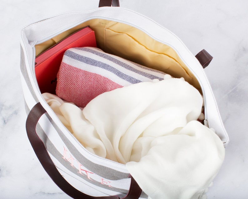 May include: A striped canvas tote bag with brown leather handles. The bag is filled with a white blanket, a red and white striped towel, and a red notebook.
