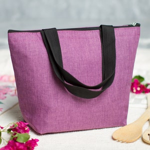 May include: A purple insulated tote bag with black handles and a zipper closure. The bag is sitting on a white surface with pink flowers and wooden utensils.