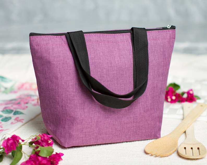 May include: A purple insulated tote bag with black handles and a zipper closure. The bag is sitting on a white surface with pink flowers and wooden utensils.