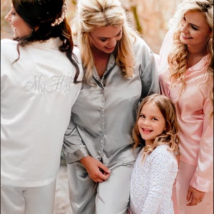 May include: Four women and a young girl wearing matching pajamas in a forest setting. The women are wearing gray and pink satin pajamas, while the girl is wearing a white and blue floral pajama set. The bride-to-be is wearing a white satin robe with the text "Mrs. Hill" embroidered on the back.