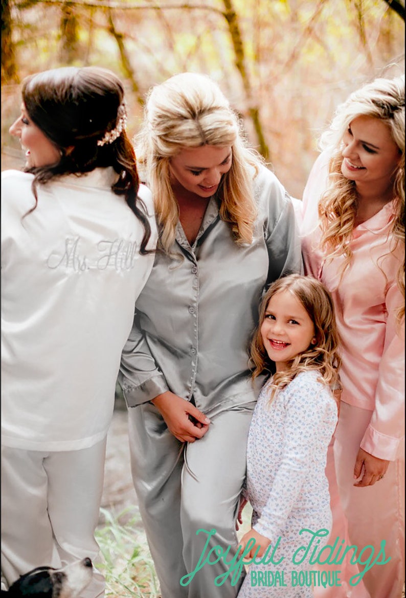 May include: Four women and a young girl wearing matching pajamas in a forest setting. The women are wearing gray and pink satin pajamas, while the girl is wearing a white and blue floral pajama set. The bride-to-be is wearing a white satin robe with the text "Mrs. Hill" embroidered on the back.