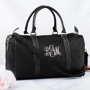 以下が含まれることがあります： ピンクのモノグラムが「B＆M」と刺繍された黒いダッフルバッグ。