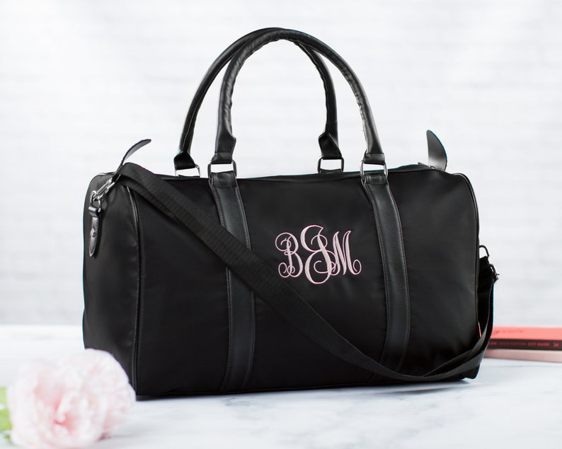 以下が含まれることがあります： ピンクのモノグラムが「B＆M」と刺繍された黒いダッフルバッグ。