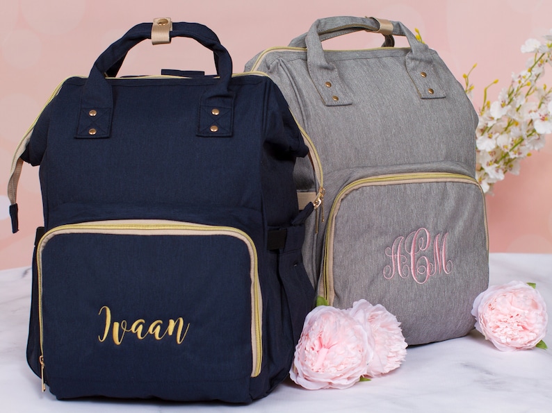 May include: Two personalized diaper bags, one navy blue with the name "Ivaan" embroidered in gold thread and one gray with the monogram "ACM" embroidered in pink thread.