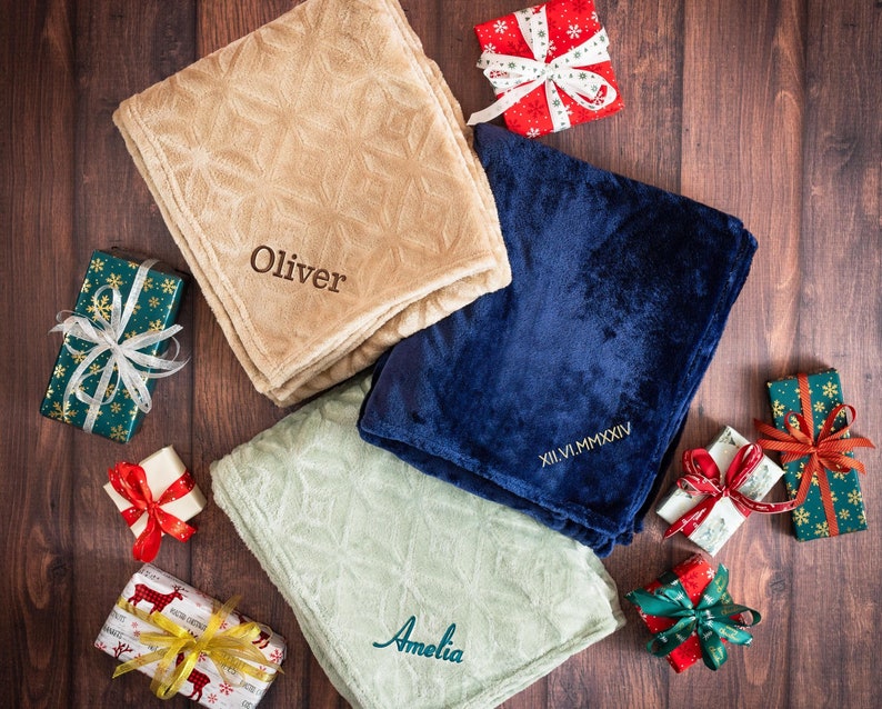 May include: Three plush blankets with personalized embroidery. The blankets are beige, blue, and green. The beige blanket has the name "Oliver" embroidered on it. The blue blanket has the Roman numeral date "XII.VI.MMMXIV" embroidered on it. The green blanket has the name "Amelia" embroidered on it. The blankets are surrounded by wrapped gifts with red and green ribbon.