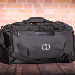 K&ouml;nnte beinhalten: Schwarze Reisetasche mit Rei&szlig;verschluss und Schultergurt. Die Tasche hat ein graues gesticktes Monogramm "CD" auf der Vorderseite.