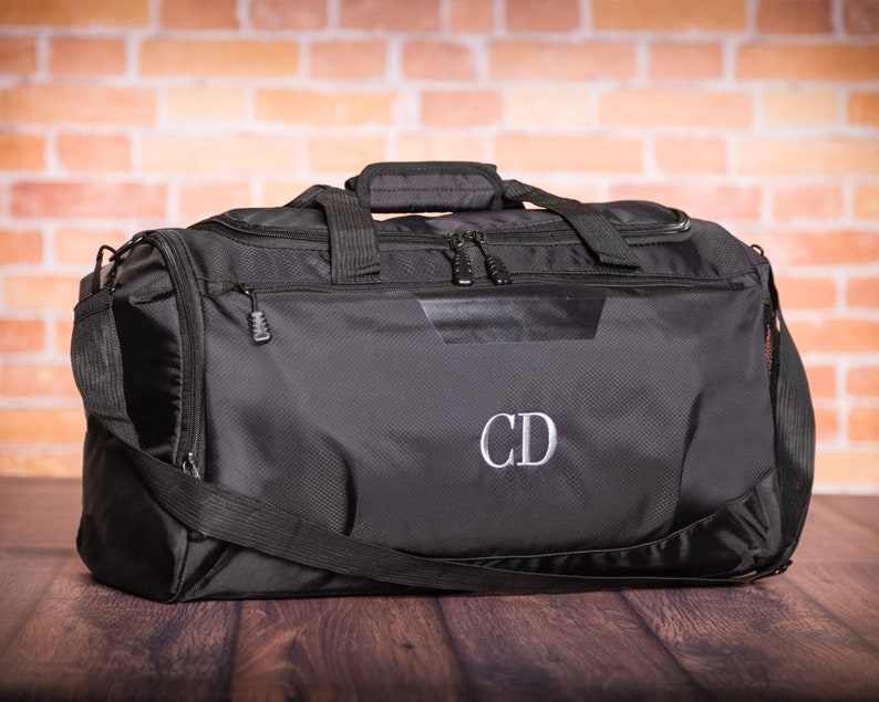 K&ouml;nnte beinhalten: Schwarze Reisetasche mit Rei&szlig;verschluss und Schultergurt. Die Tasche hat ein graues gesticktes Monogramm "CD" auf der Vorderseite.