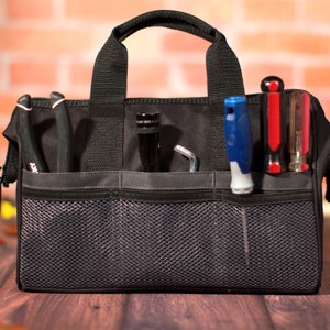 K&ouml;nnte beinhalten: Eine schwarze Werkzeugtasche mit mehreren Taschen und einem Netzfach. Die Tasche ist mit verschiedenen Werkzeugen gef&uuml;llt, darunter Schraubendreher, Zangen und ein Schraubenschl&uuml;ssel.