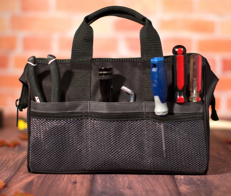 K&ouml;nnte beinhalten: Eine schwarze Werkzeugtasche mit mehreren Taschen und einem Netzfach. Die Tasche ist mit verschiedenen Werkzeugen gef&uuml;llt, darunter Schraubendreher, Zangen und ein Schraubenschl&uuml;ssel.