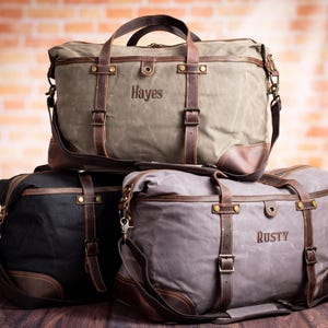 May include: Three canvas duffel bags in olive green, black, and gray, stacked on top of each other. Each bag features leather accents, straps, and handles. The olive green bag has "Hayes" printed on it, and the gray bag has "Rusty" printed on it.