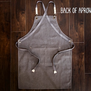May include: Back view of a gray canvas apron with adjustable straps and a large pocket. The straps are crossed in the back and have brass hardware. The text "BACK OF APRON" is printed in white on the top right corner of the apron.