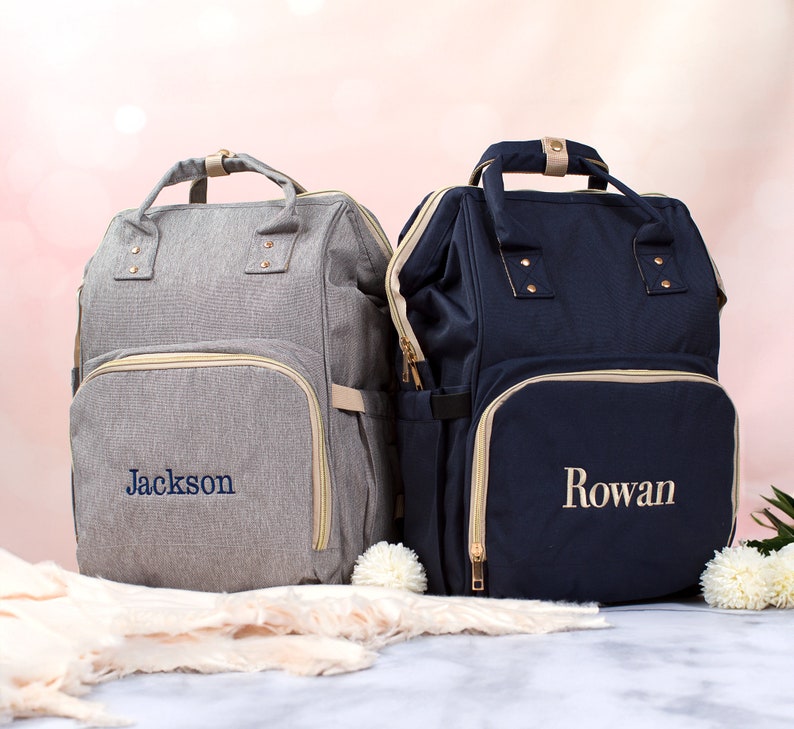 May include: Two backpacks with top handles and front zippered pockets. The left backpack is heather gray with the name Jackson embroidered in blue. The right backpack is navy blue with the name Rowan embroidered in light beige thread.