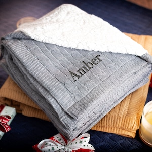 Pode incluir: Uma manta de malha cinzenta com cabo e forro de sherpa branco. A manta &eacute; personalizada com o nome "Amber" bordado em fio preto.