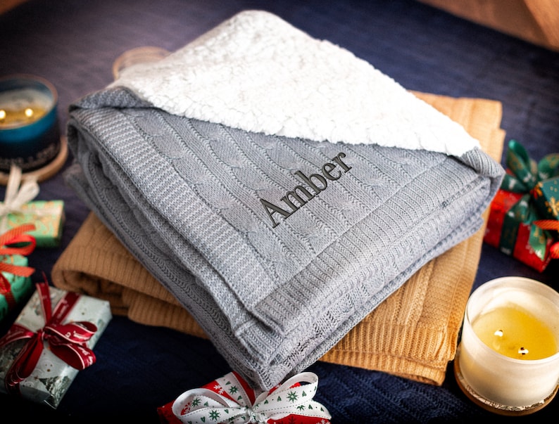 Pode incluir: Uma manta de malha cinzenta com cabo e forro de sherpa branco. A manta &eacute; personalizada com o nome "Amber" bordado em fio preto.
