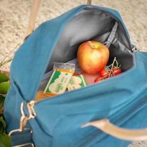 May include: A teal corduroy lunch bag with a top handle and a shoulder strap. Inside the bag are a red apple, cherry tomatoes, and two snack packs. The bag is open, revealing a gray interior lining.
