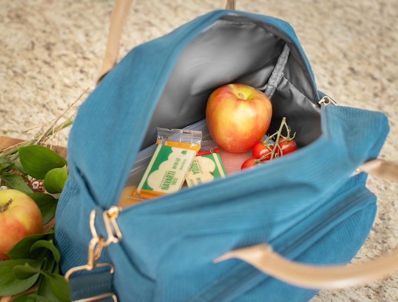 May include: A teal corduroy lunch bag with a top handle and a shoulder strap. Inside the bag are a red apple, cherry tomatoes, and two snack packs. The bag is open, revealing a gray interior lining.