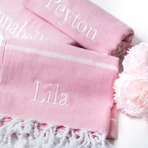 Puede incluir: Tres toallas turcas rosas con flecos blancos. Las toallas est&aacute;n bordadas con los nombres "Annabelle", "Peyton" y "Lila".