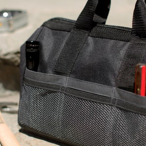 Puede incluir: Una bolsa de herramientas negra con m&uacute;ltiples bolsillos y un compartimento de malla. La bolsa tiene una cremallera y dos asas. La bolsa est&aacute; llena de herramientas, incluyendo una linterna y un destornillador.