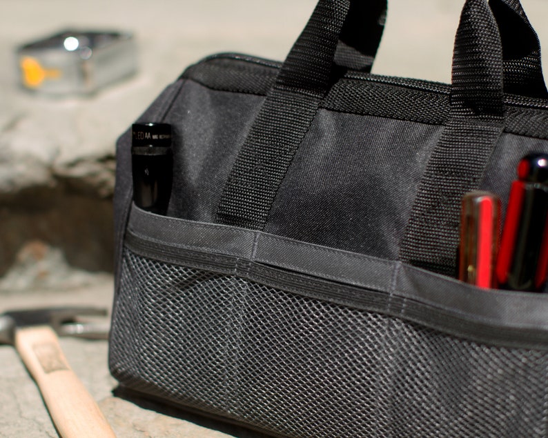 Puede incluir: Una bolsa de herramientas negra con m&uacute;ltiples bolsillos y un compartimento de malla. La bolsa tiene una cremallera y dos asas. La bolsa est&aacute; llena de herramientas, incluyendo una linterna y un destornillador.
