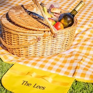 Puede incluir: Una cesta de picnic de mimbre con tapa cerrada, llena de comida y una botella de vino, se encuentra sobre una manta a cuadros amarillos y blancos. Una alfombra amarilla con el texto "The Lees" est&aacute; en primer plano.