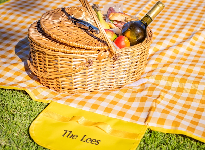 Puede incluir: Una cesta de picnic de mimbre con tapa cerrada, llena de comida y una botella de vino, se encuentra sobre una manta a cuadros amarillos y blancos. Una alfombra amarilla con el texto "The Lees" est&aacute; en primer plano.