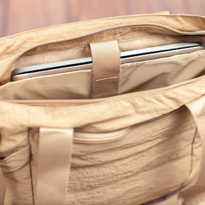 May include: A beige tote bag with a laptop inside. The bag has a zippered main compartment and a shoulder strap. The laptop is secured with a fabric strap. The bag is made of a crinkled fabric.