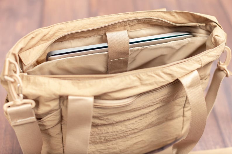 May include: A beige tote bag with a laptop inside. The bag has a zippered main compartment and a shoulder strap. The laptop is secured with a fabric strap. The bag is made of a crinkled fabric.