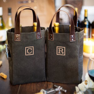 May include: Two dark gray canvas wine totes with brown leather handles. The totes have a pocket on the side and are monogrammed with the letters "C" and "R".