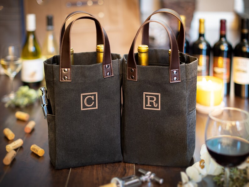 May include: Two dark gray canvas wine totes with brown leather handles. The totes have a pocket on the side and are monogrammed with the letters "C" and "R".