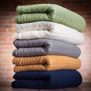 May include: A stack of folded cable-knit blankets in various colors, including green, white, gray, mustard yellow, and navy blue. The blankets are neatly arranged, showcasing their textured design and soft appearance. The background is a brick wall.