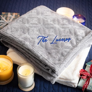 May include: A gray and white plush blanket with a diamond pattern and the text "The Luceros" embroidered on it. The blanket is folded and placed on a blue knitted surface with candles and wrapped gifts.
