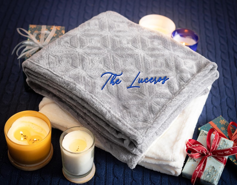 May include: A gray and white plush blanket with a diamond pattern and the text "The Luceros" embroidered on it. The blanket is folded and placed on a blue knitted surface with candles and wrapped gifts.