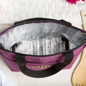 May include: Purple insulated lunch bag with black handles and a gold monogram. The bag is open and the silver lining is visible.