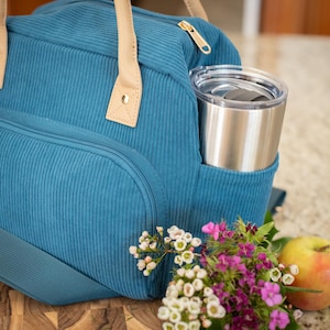 May include: Teal corduroy lunch bag with tan leather handles and a gold zipper. A stainless steel tumbler sits in the side pocket. A small bouquet of flowers and a yellow apple are next to the bag.